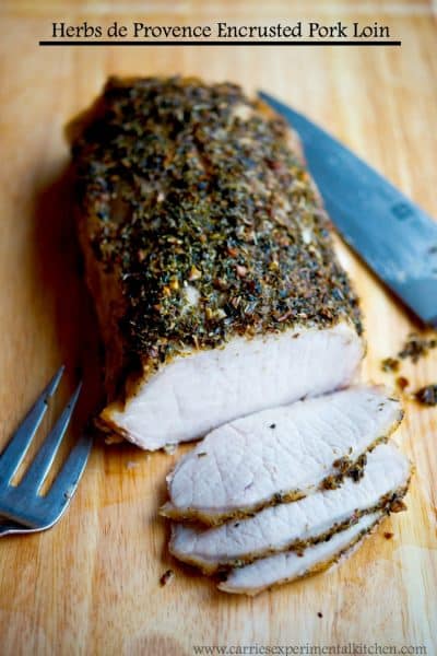 A close up of sliced Herbs de Provence Encrusted Pork Loin on a wooden cutting board.