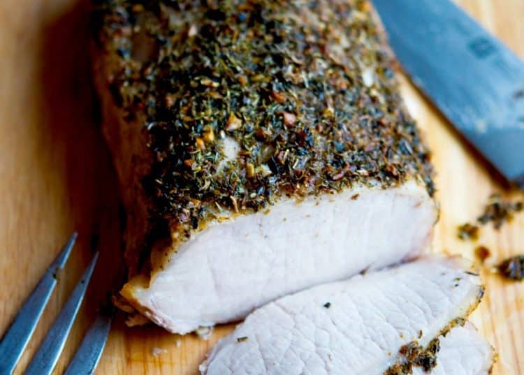 A close up of sliced Herbs de Provence Encrusted Pork Loin on a wooden cutting board.