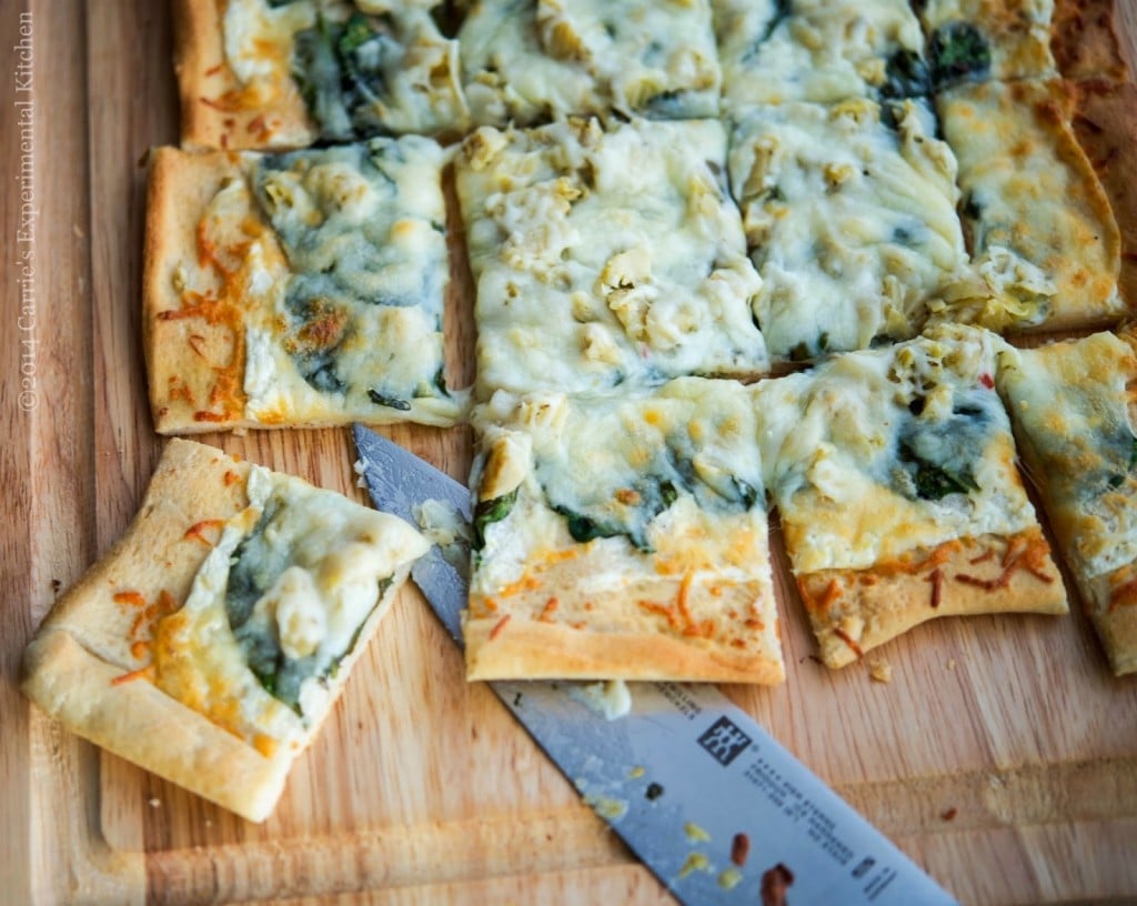 Spinach artichoke flatbread pizza cut into squares on a wooden cutting board. 