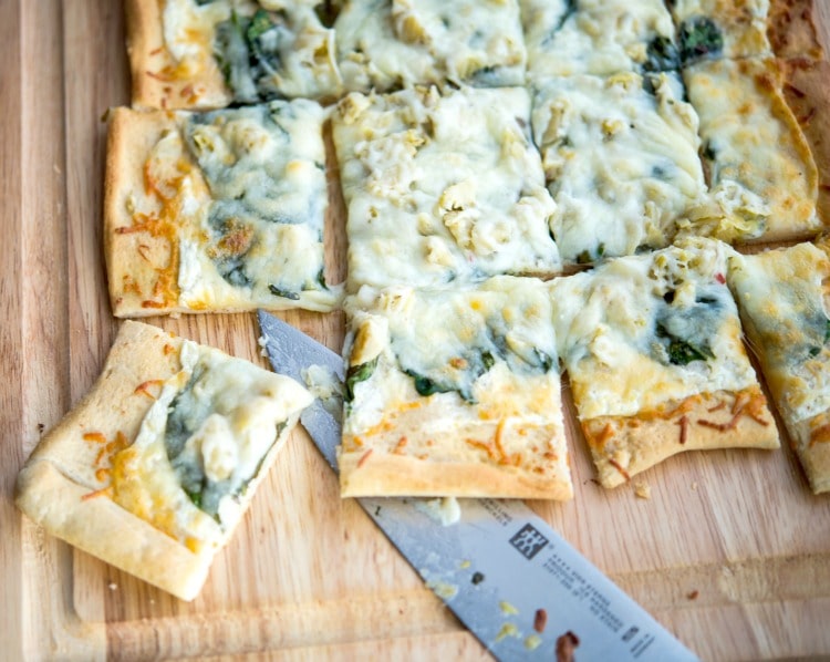 Spinach and Artichoke Flatbread cut into squares on a wooden cutting board. 