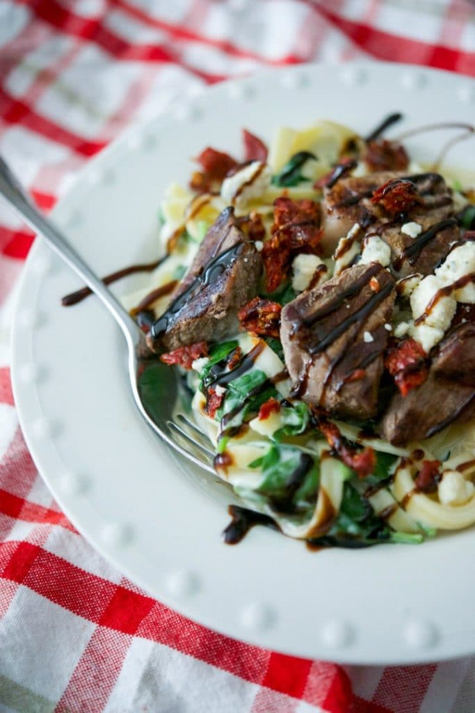 Grilled steak over fettuccine alfredo tossed with fresh spinach and Gorgonzola cheese; then topped with sun dried tomatoes and balsamic drizzle in a bowl with a fork. 