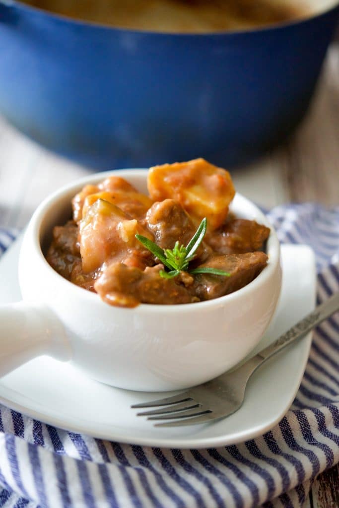 Beef stew made with zucchini, garlic, onions, fire roasted tomatoes, fresh rosemary, horseradish and red wine in a white crock. 