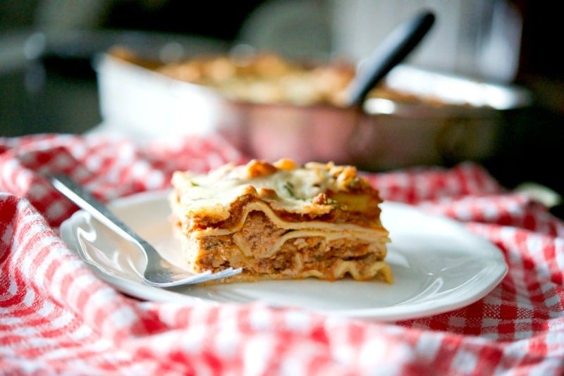 A piece of lasagna on a white plate with a fork and a tray in the background. 