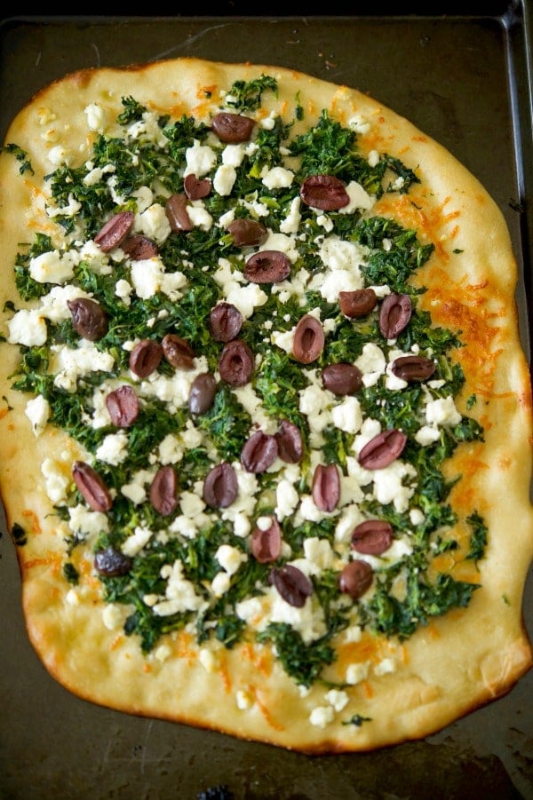 Flatbread topped with spinach, crumbled Goat cheese and Kalamata olives on a cutting board. 