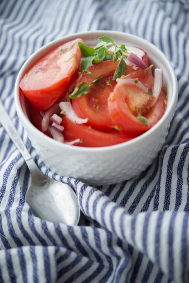 Sliced tomatoes in a white bowl on top of a blue and white napkin with a spoon on the side. 