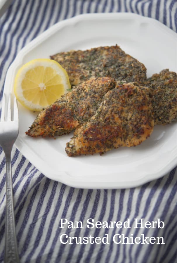 Pan fried boneless chicken breasts dredged in herbs and spices on a plate with a halved lemon. 