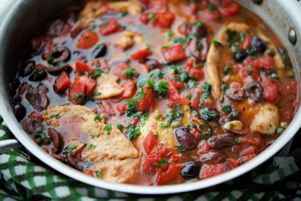 a close up of chicken in a skillet with olives, tomatoes and capers