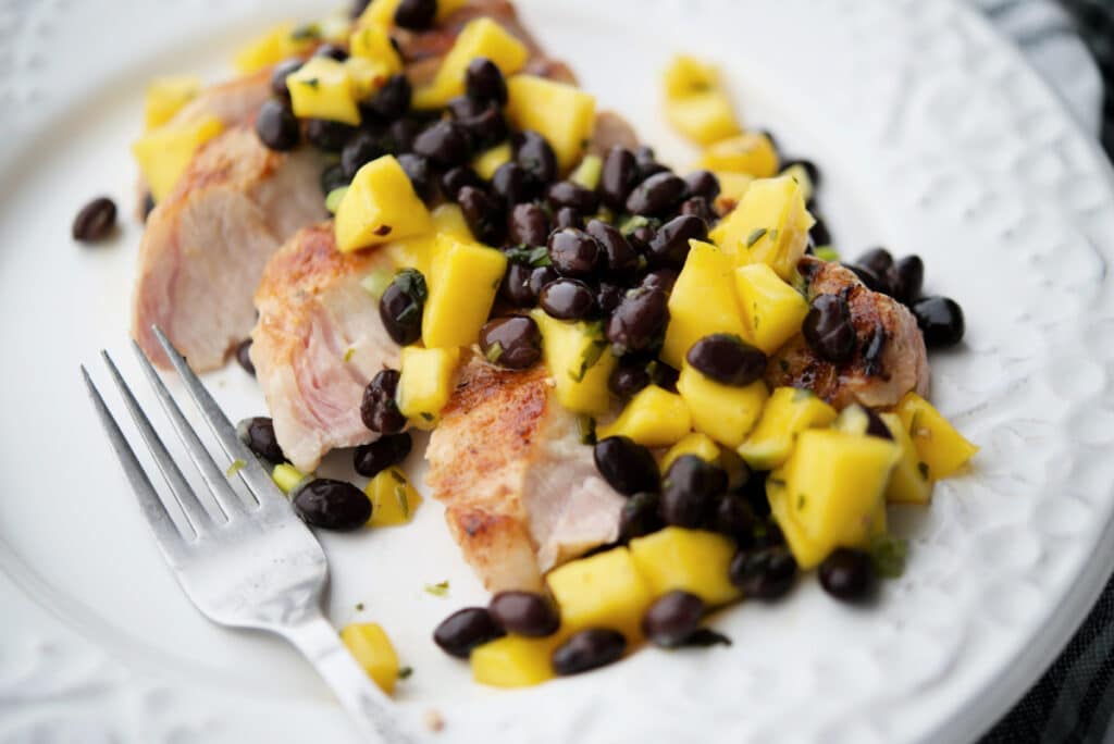 a close up of slice grilled pork chop on a white plate with mango black bean salsa